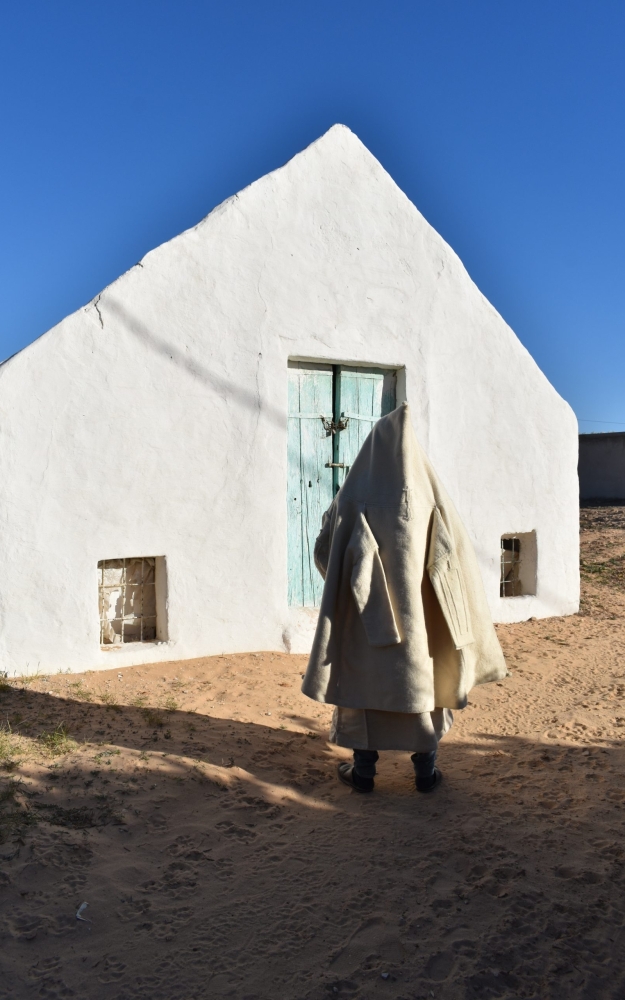 A man wearing traditional clothing in front of a weaving shop | صورة بلباس تقليدي رجالي امام حانوت نسيج حياكة